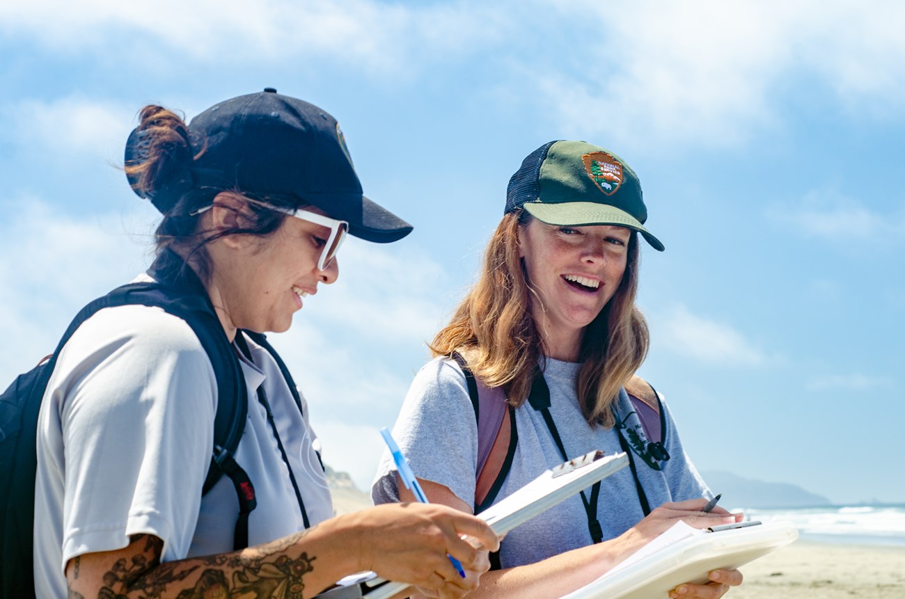 Rachel on Ocean Beach, smiling as she converses with a wildlife intern. Both carry clipboards for recording western snowy plover data.