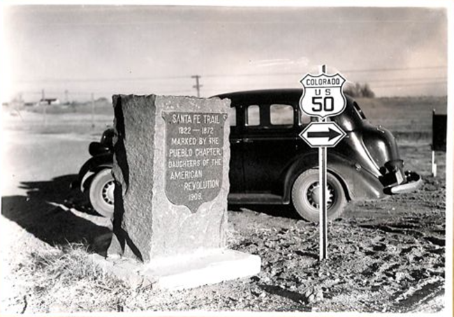 Black 1930s vehicle parks by a Santa Fe Trail marker and sign for Colorado Hwy 50