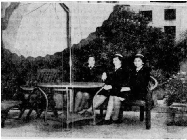 Grainy photo of three young women sitting at a table with an umbrella. They wear dark suits and caps. They are sitting casually.