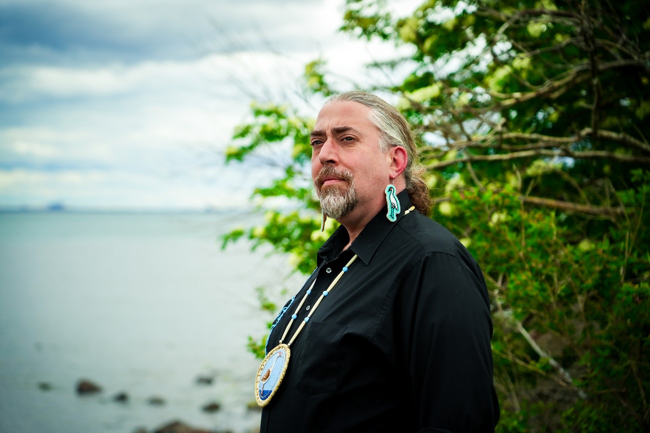 Headshot of a person wearing beaded earrings and a medallion with crane motif in front of a lake.