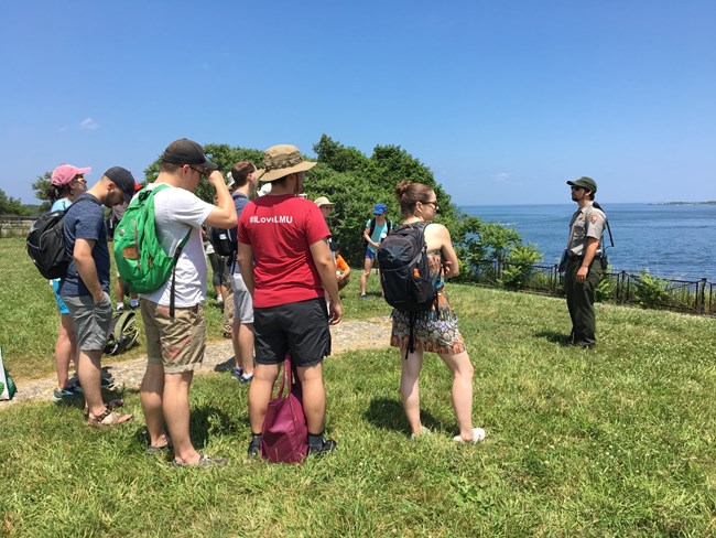 A ranger giving a talk on grass with a harbor in the background.