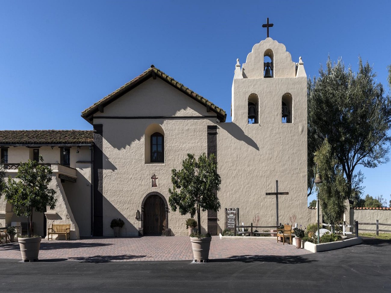 Tan building, shorter on the left, taller in the middle, and a bell tower to the right. Roof and doors are brown. There are trees in the foreground.