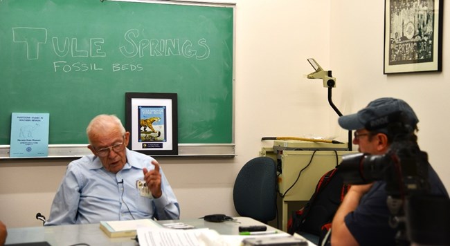 Photograph of 2 people seated at a table. Chalboard behind the interviewee says "Tule Springs Fossil Beds"
