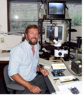Photograph of a man sitting in front of monitors and scientific equipment such as microscope.