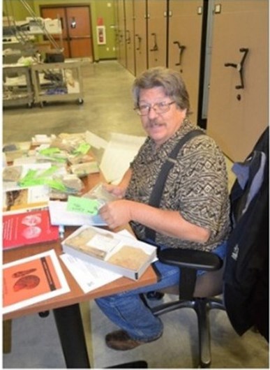 Photograph of a man sitting in a chair at a table with fossils and publications. Museum cabinets are in the background.