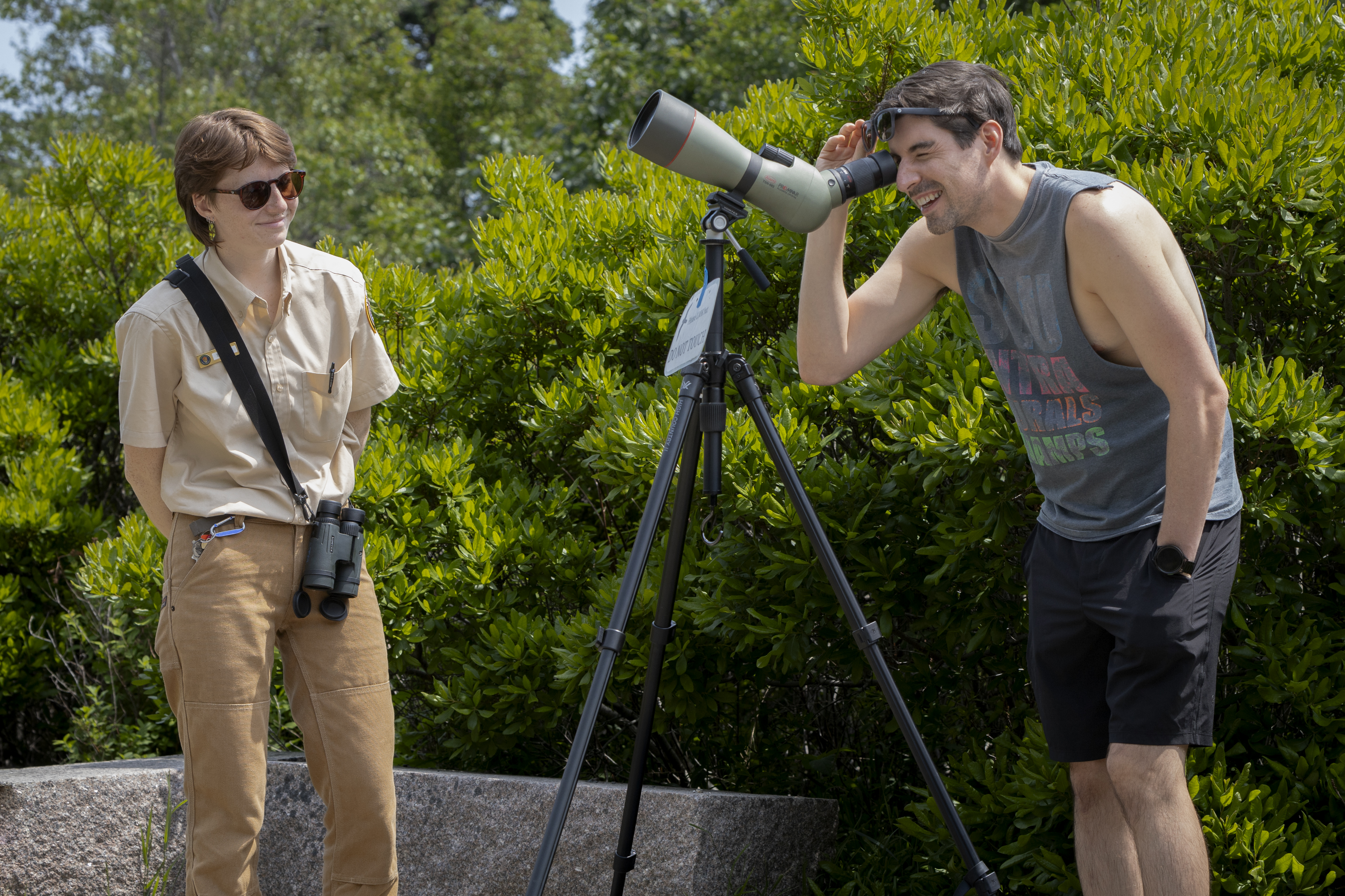 Intern talking to a visitor who is looking through a spotting scope pointed at a nesting peregrine falcon chick on the Precipice Trail.