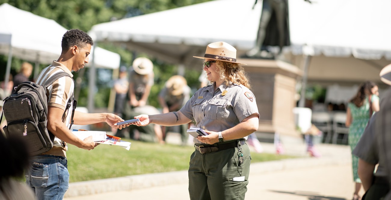 A woman handing a National Park Service passport to a visitor with a statue in the background.