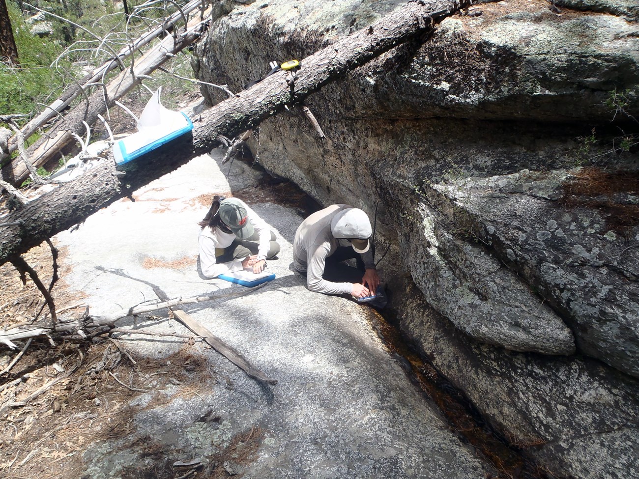 Two people sitting on a very large boulder collecting data next to a narrow channel of water passing between large rocks