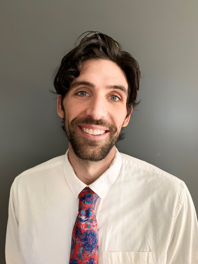 Headshot of a white man with brown hair and a beard wearin a white shirt and a floral tie