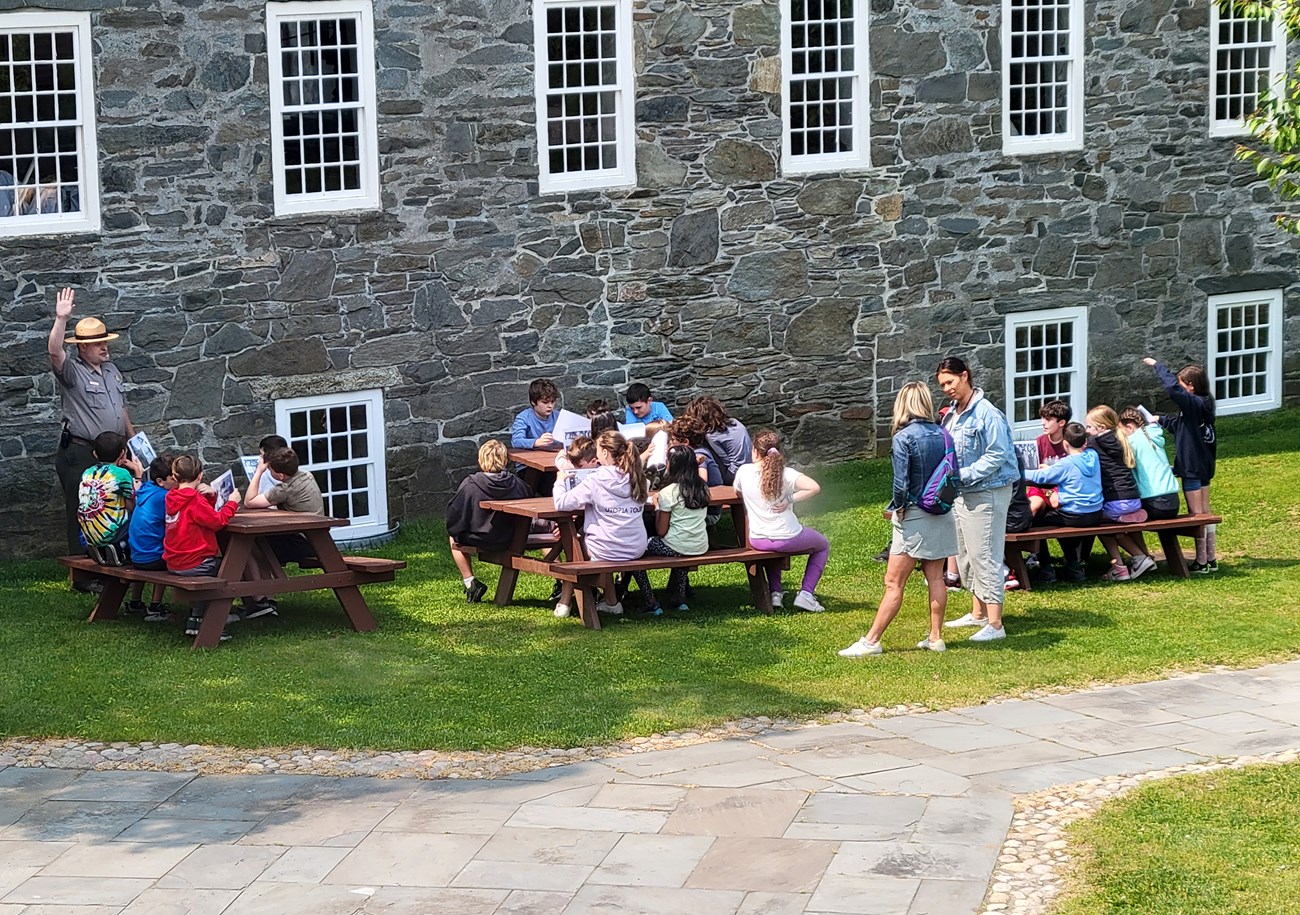 NPS ranger with a whistle holds his hand up in front of schoolchildren seated at picnic tables outside a gray stone building with white windows