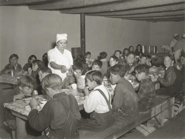 Black and white photo of young kids eating at long lunch tables. A lunch lady in a white smock and hair-covering gestures toward one of the kids. They are in windowless room with a low ceiling and exposed beams.