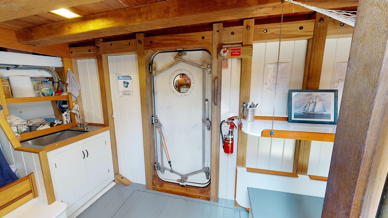 Below deck on the schooner, facing a door and a sink. Looking closely through the door's small round window, we see a person peering at the camera.