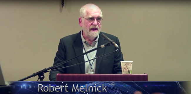 Robert Melnick speaking at lectern. This image links to a non-audio described YouTube presentation.