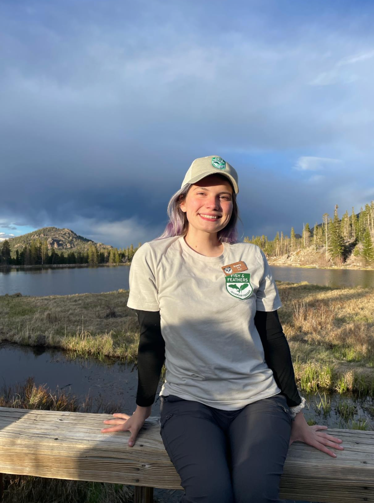 A woman sitting on a bench with water, trees, and mountains behind her