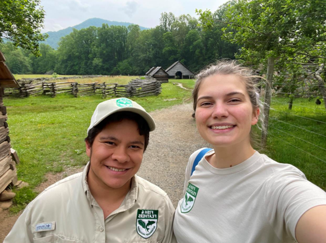 Two female individuals taking a selfie together with wooden fencing, small wooden houses, and trees in the background
