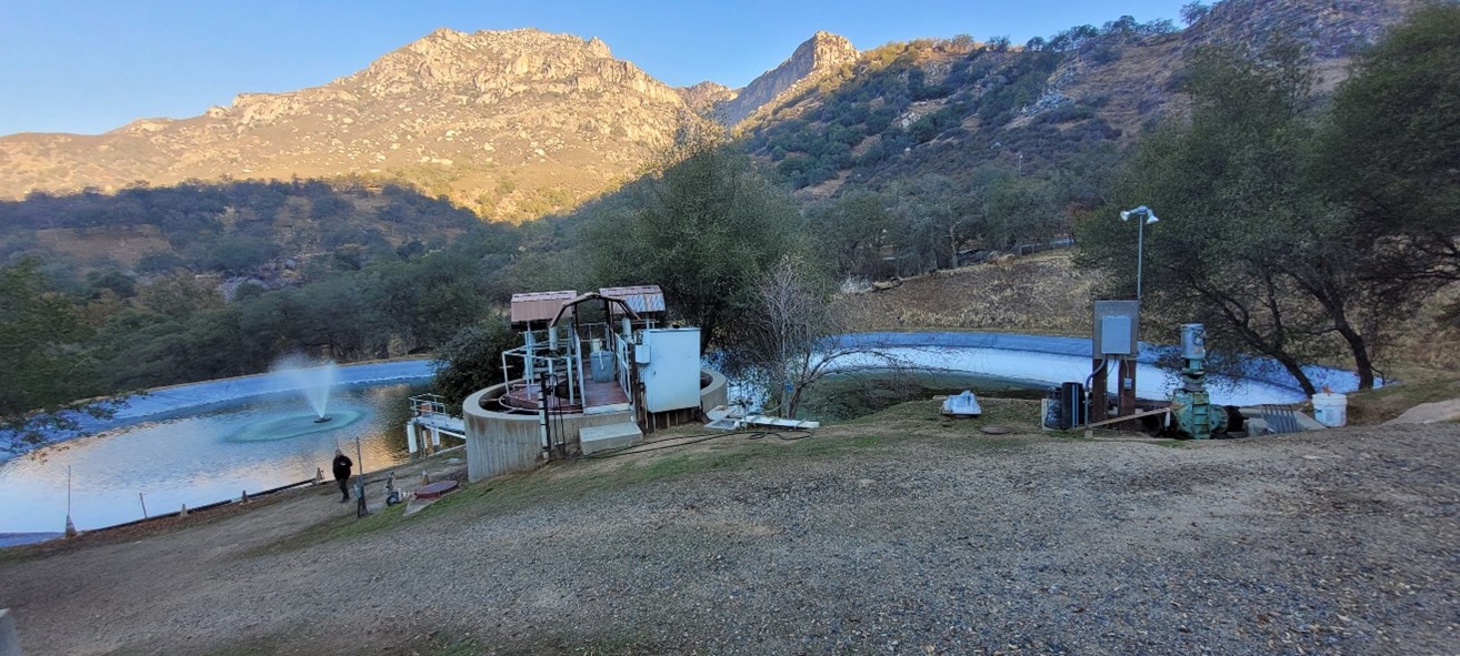 An out-of-date water pump system sits in front of a man-made water basin with black lining. A mountain range sits behind it.
