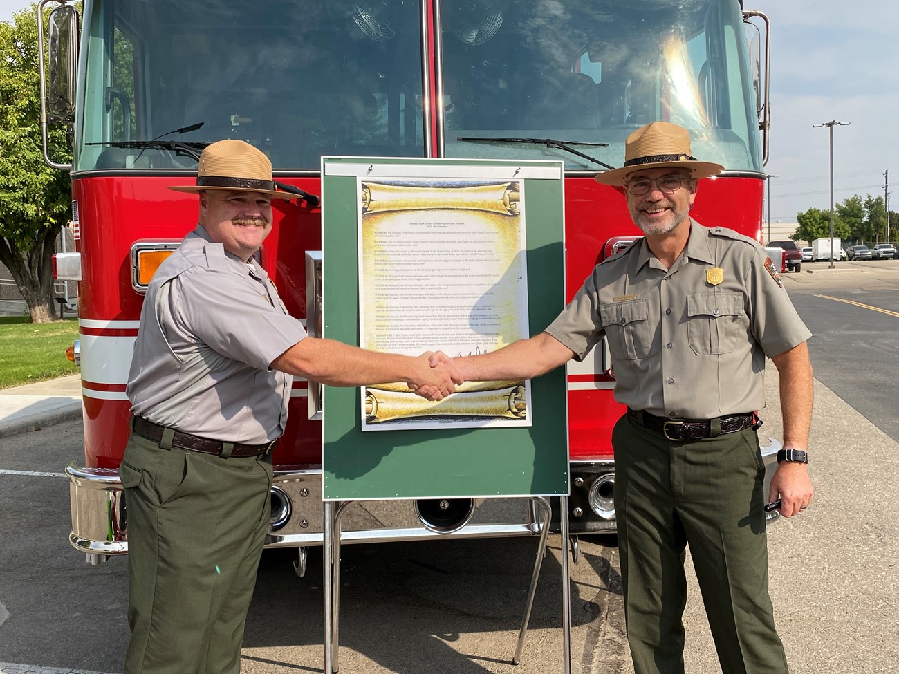 Two people shake hands in front of a fire engine with a board holding a sign.