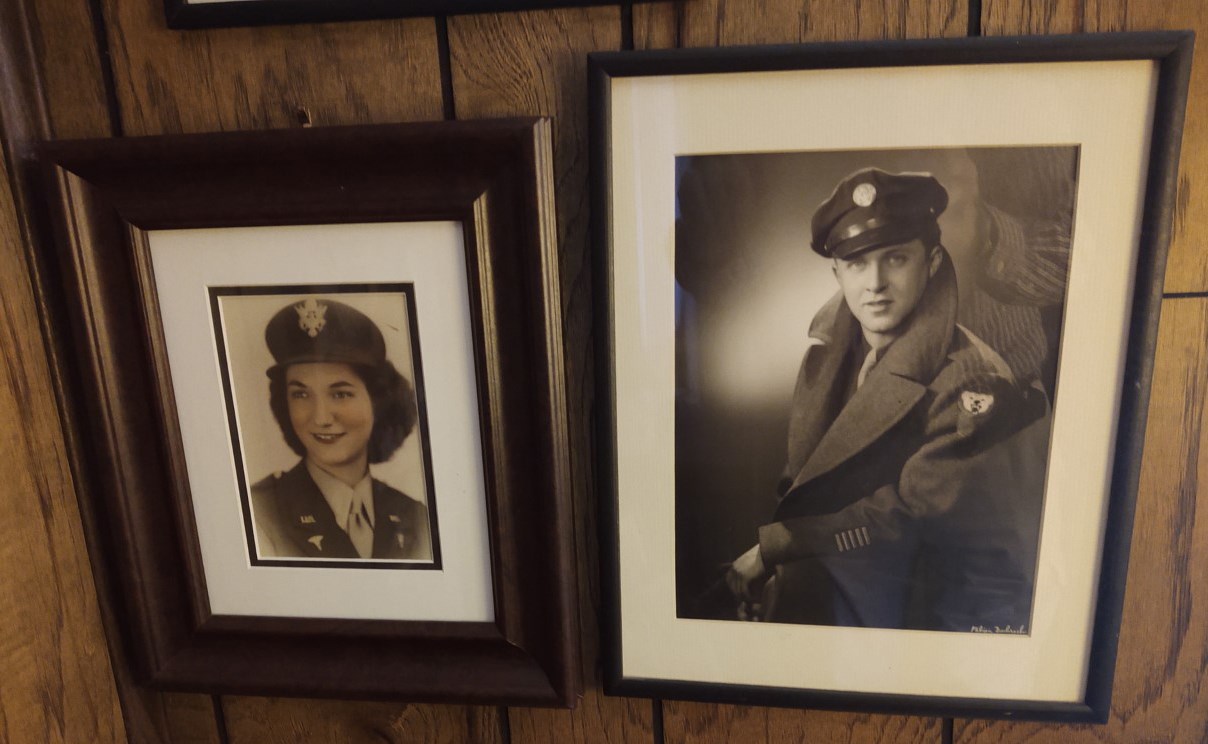 An image of two framed photos hanging on a wall. The photo on the left is of a woman smiling, in a military uniform. The photo on the right is of a man, also in a military uniform.