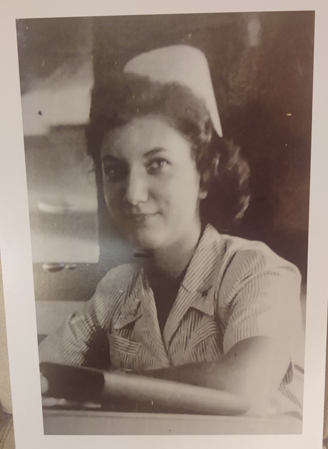 A black and white photo of a woman smiling. She is in a nursing uniform with thin stripes and wearing a white cap. Her hair is brown, curly and shoulder length.