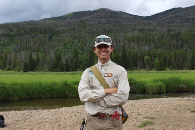 A young individual posing in front of green lush mountains, wearing a hat, with arms crossed.