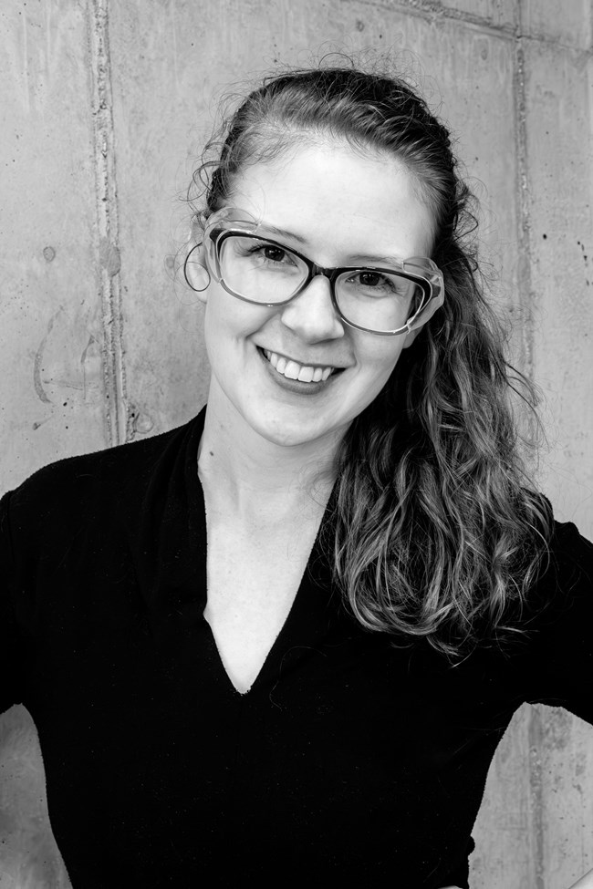 Smiling woman wearing glasses wearing a dark blouse with long wavy hair pulled back and over one shoulder.
