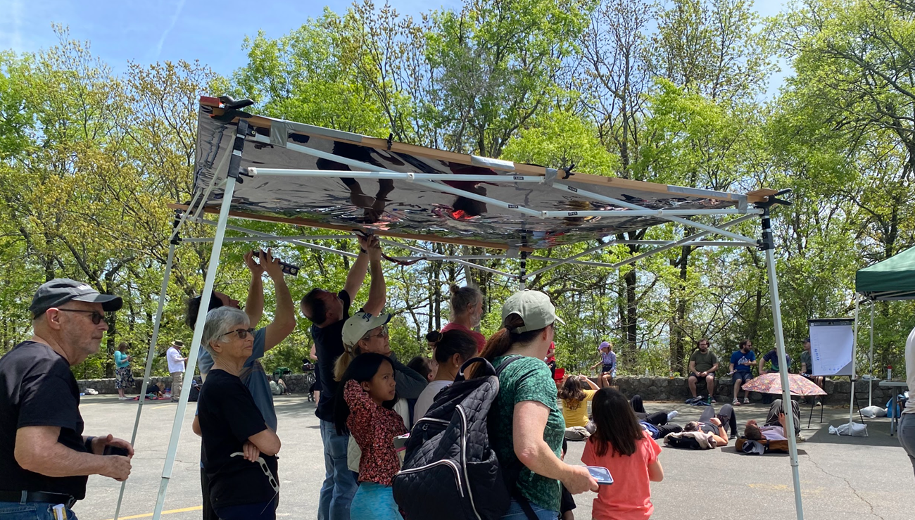 A canopy made of blacked out eclipse viewing material allows visitors to observe the sun safely without holding up special glasses. A small gorup gathers inside and looks up.