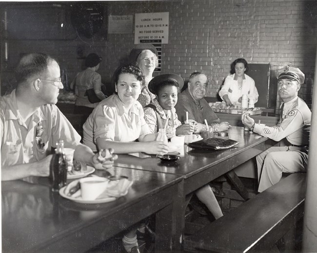 Black and white photo of mixed race, mixed gender group eating lunch at a long table. Man on far right is wearing a military uniform. Woman cleaning dishes in background.