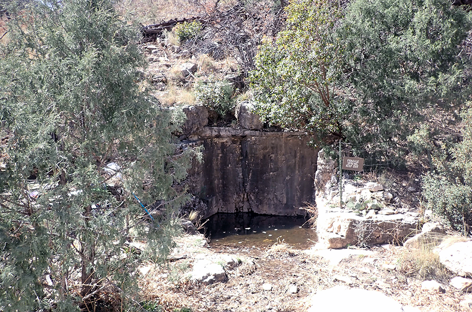 A spring bounded by three carved vertical rock walls. A fence on the perimeter has a sign that reads “FRAGILE SPRING KEEP OUT”.