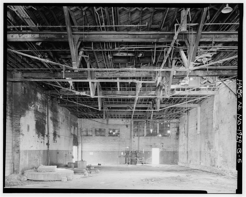 Black and white photo of a rundown building interior, with exposed piping along the ceiling.