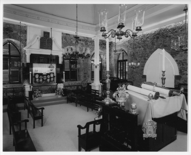 Interior of synagogue with raised Ark on far wall, holding six Torahs, and the Bimah (or pulpit) against the west wall facing the Ark. Center square area defined by four Ionic columns resting on tall pedestals. Raised platforms on the south and north side