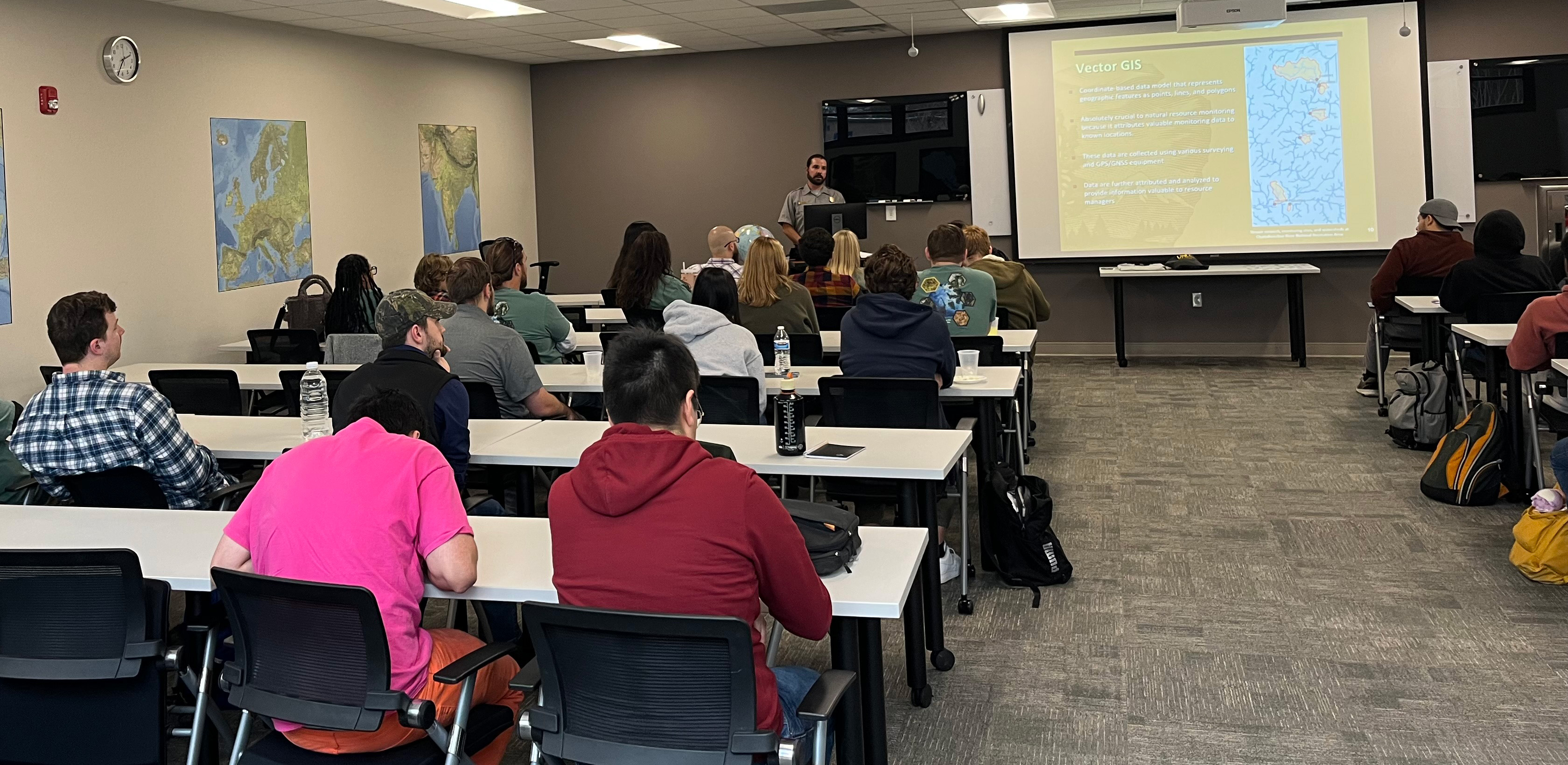 Man in uniform talking to people sitting at desks.