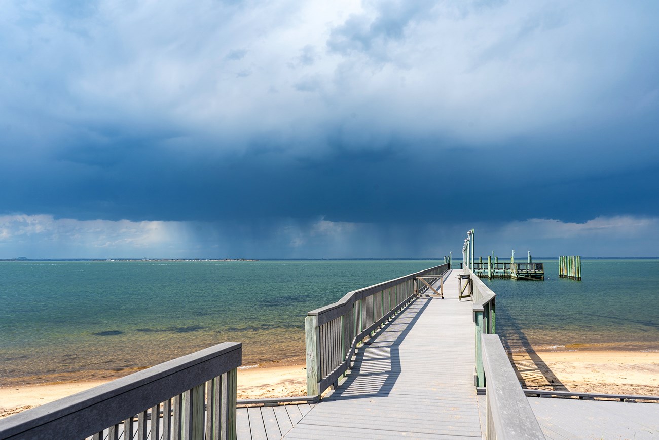 A photo of a gray dock stretching out into blue-green water, with a storm approaching.