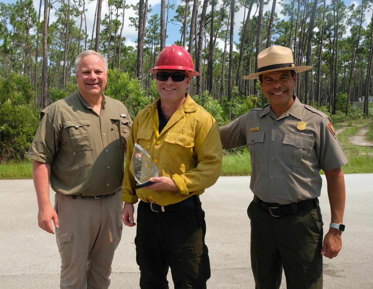 Three men pose standing on asphalt in front of a forest. The man in center is holding an award.