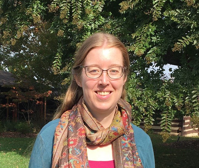 a woman with straight brown hair and glasses stands in the sunshine under a tree.