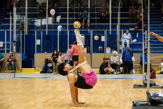 a young man uses two hands to stabilize his body on the ground while he reaches up with both feet to kick a dangling ball.
