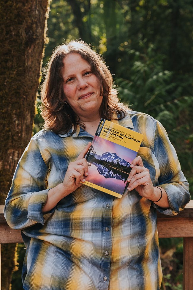 Syren Nagakyrie stands outside smiling in front of a forest holding a copy of The Disabled Hiker’s Guide to Western Washington and Oregon.