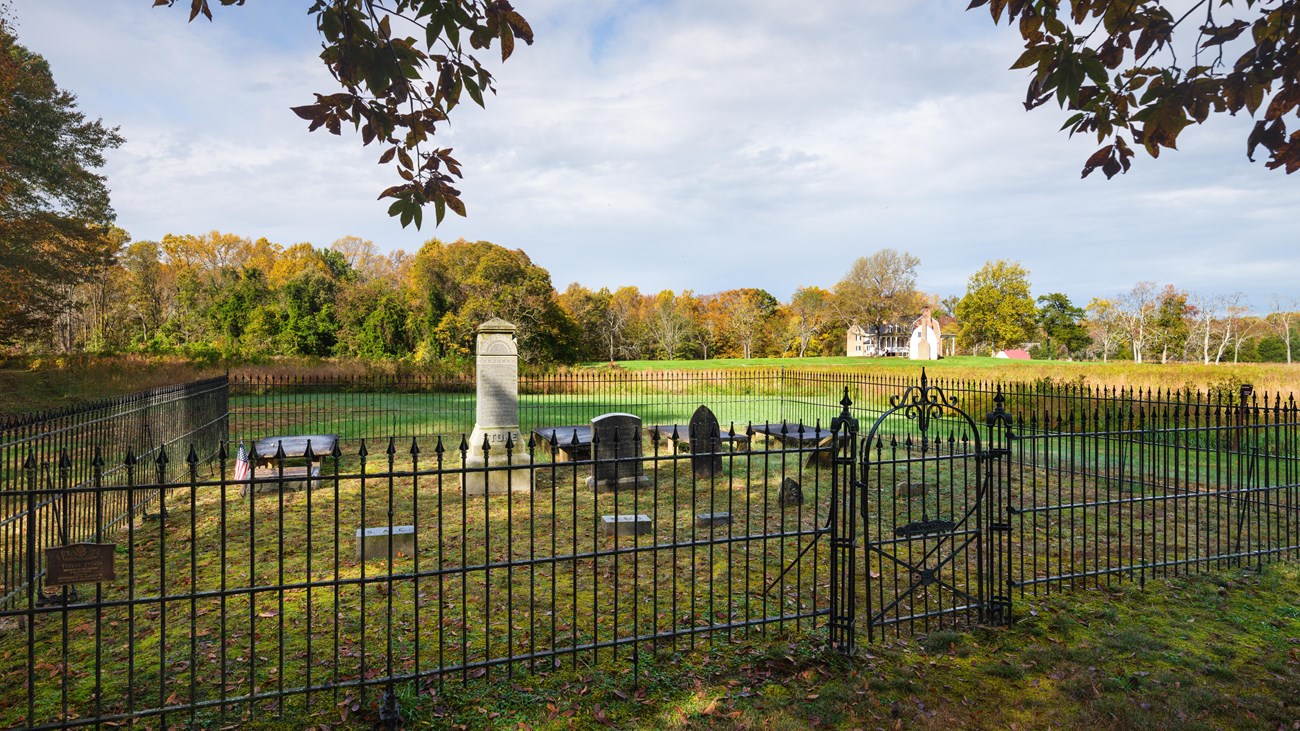 Under a gray sky, gravestones are surrounded by a black wrought iron fence, a large brick farmhouse sits in a field of green grass and trees in the background.