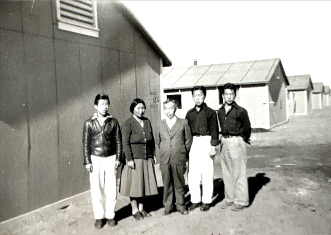 Black and white photo of a family of five (four men, one woman) standing in front of a row of barracks.