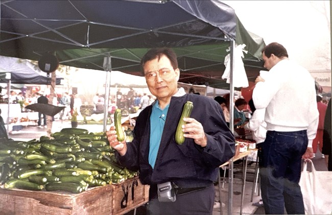 A man standing next to a produce stand holding two zuchinni.