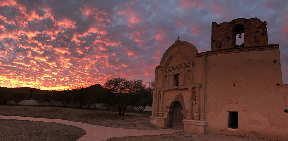 Surreal lighting on an old mission church under a mesmerizing sunset of brilliant yellow, orange, pink, and bluish clouds filling the sky.