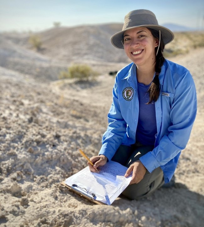 A scientists in a blue button-up shirt and a brimmed hat sits on the ground with a clipboard and pencil, taking notes on her observations of the state of the paleontological resources is Tule Springs Fossil Beds National Monument.