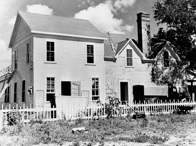 A two-story frame and stone house with construction materials and 1950s pickup truck in the yard.