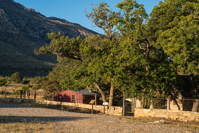 across a gravel drive a stone wall defines a ranging compound with trees, a red frame building, and a stone house.