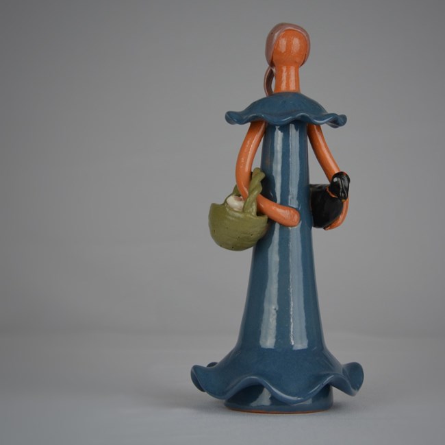 Ceramic faceless doll wearing a blue dress holding a green basket and black chicken.