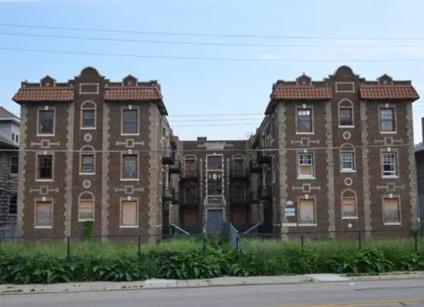 Image of a three-story brown brick apartment building with two symmetrical facades in Mediterranean Revival style. Some windows are boarded up. Overgrown greenery separates the building and a fence.