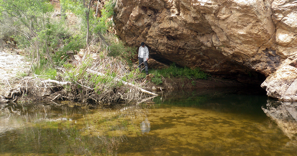 A large, shallow pool of water that extends into a cavern under a steep, large rock.