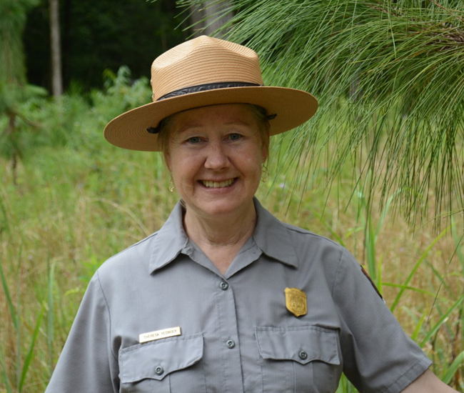 A woman in NPS uniform and flat hat smiles with green pine trees in the background