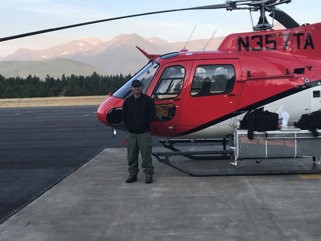Smiling man stands in front of red helicopter with mountains in the background.