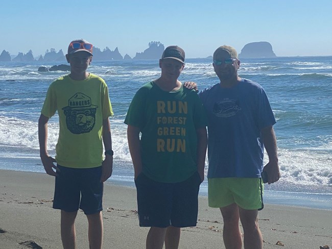 An adult male and his two sons are on a sandy beach smiling at the camera with crashing waves behind them and craggy peaks in the distance.
