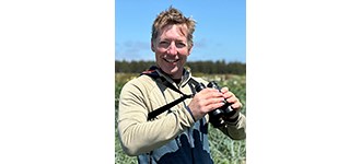 Rodhouse outside in a grassland, holding up a pair of binoculars and smiling at the camera.